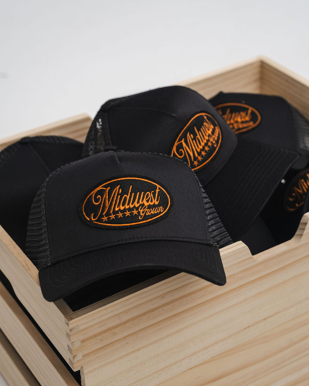 Black trucker hats with gold 'Midwest' logo in a wooden box