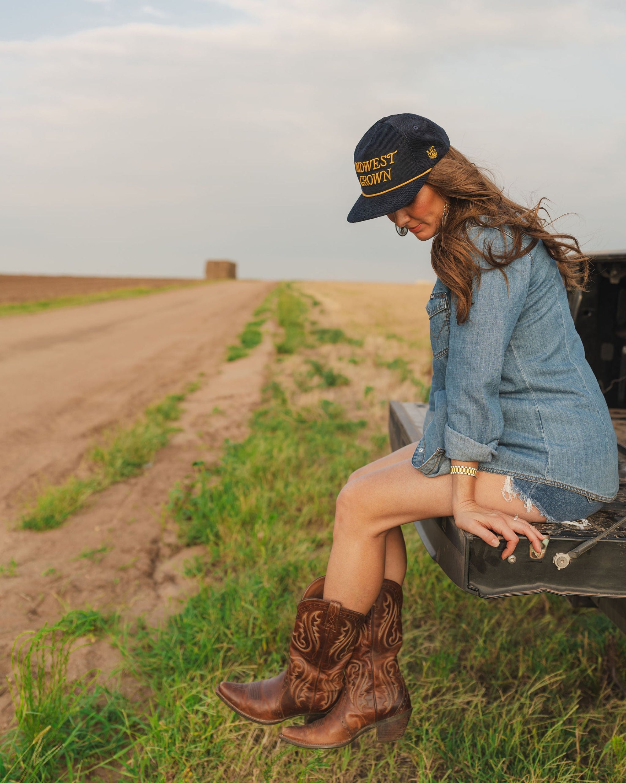Woman sitting on a truck bed in a field wearing cowboy boots, a denim jacket, and a Midwest Grown Hat.