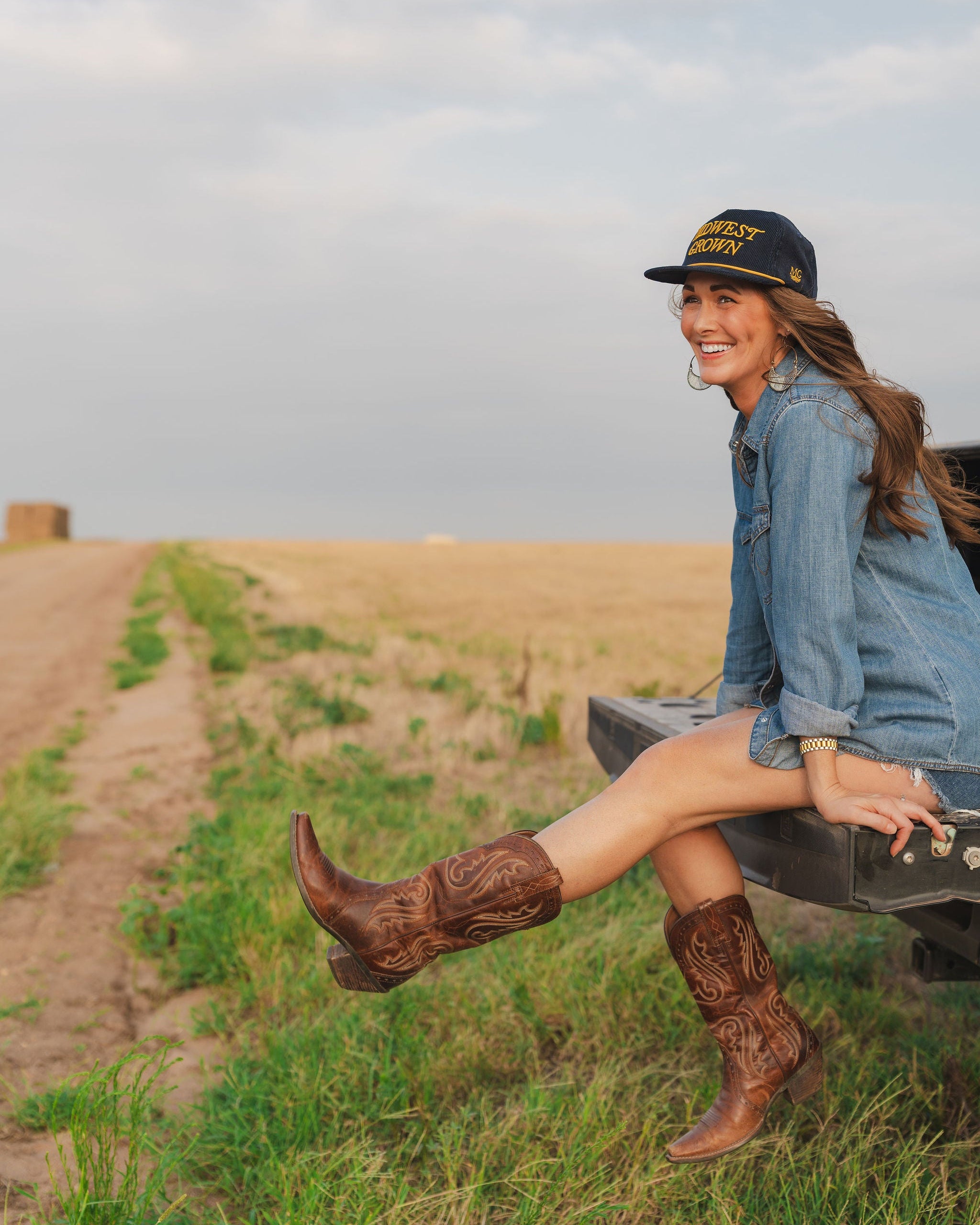 Woman in cowboy boots and denim outfit sitting on a truck bed in a field wearing a Midwest Grown Hat