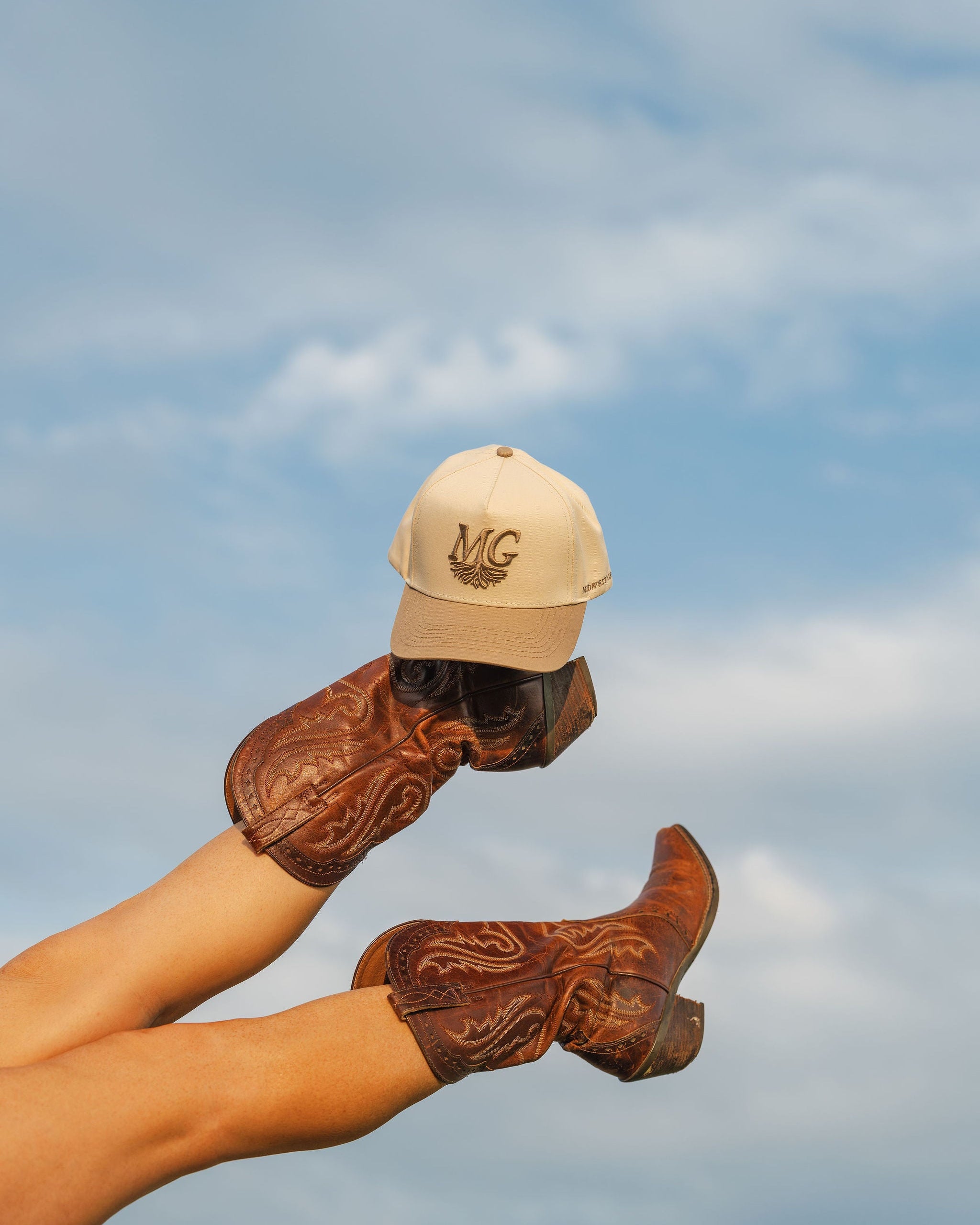 Person wearing cowboy boots and a Midwest Grown cap against a blue sky.