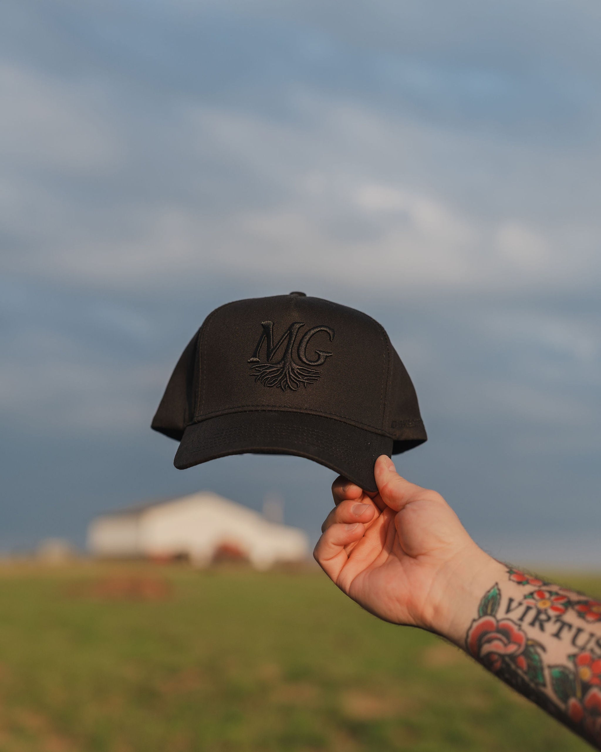 Hand holding a black cap with 'Midwest Grown' logo against a cloudy sky