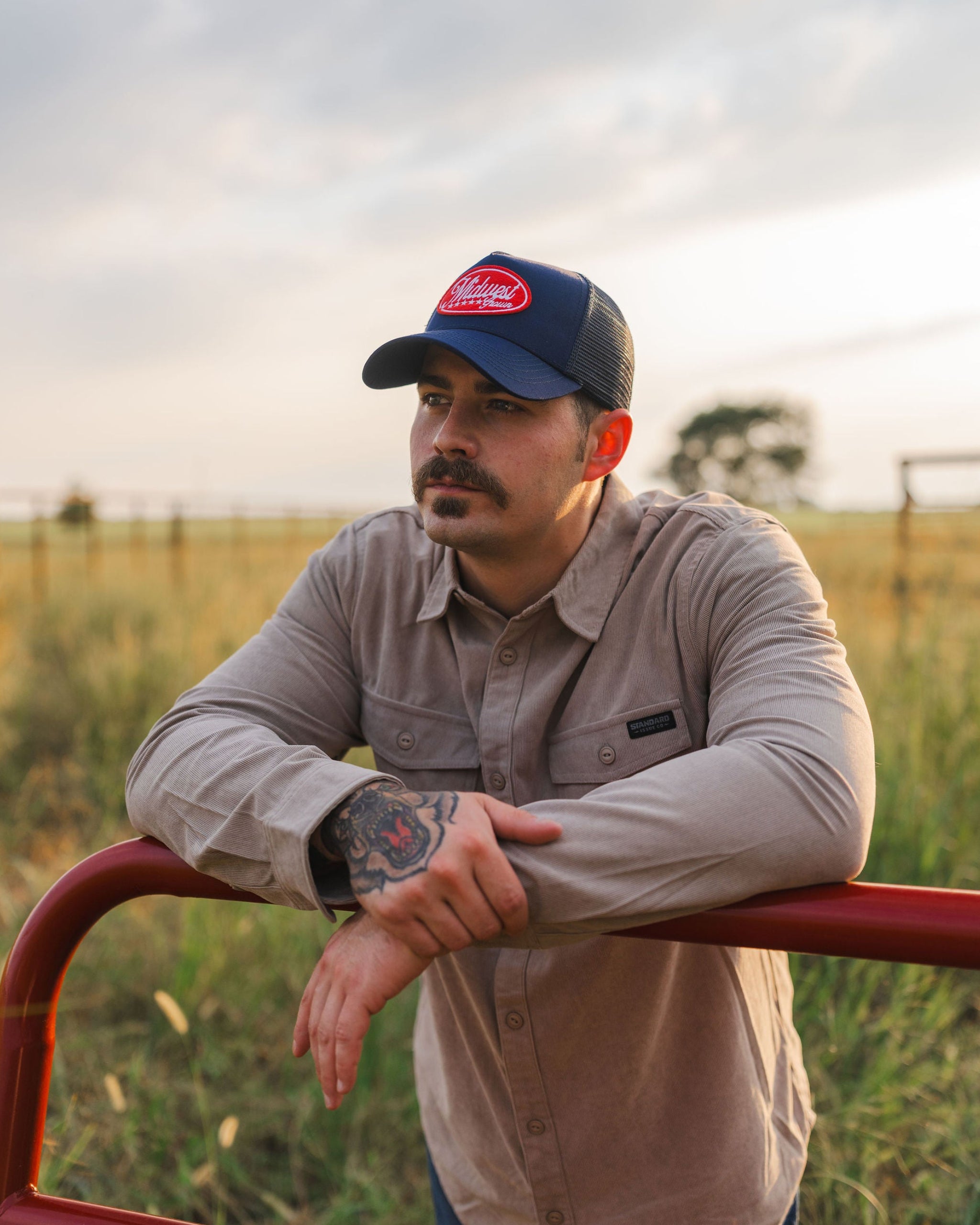 Man wearing a Midwest Grown cap and leaning on a red metal gate in a field