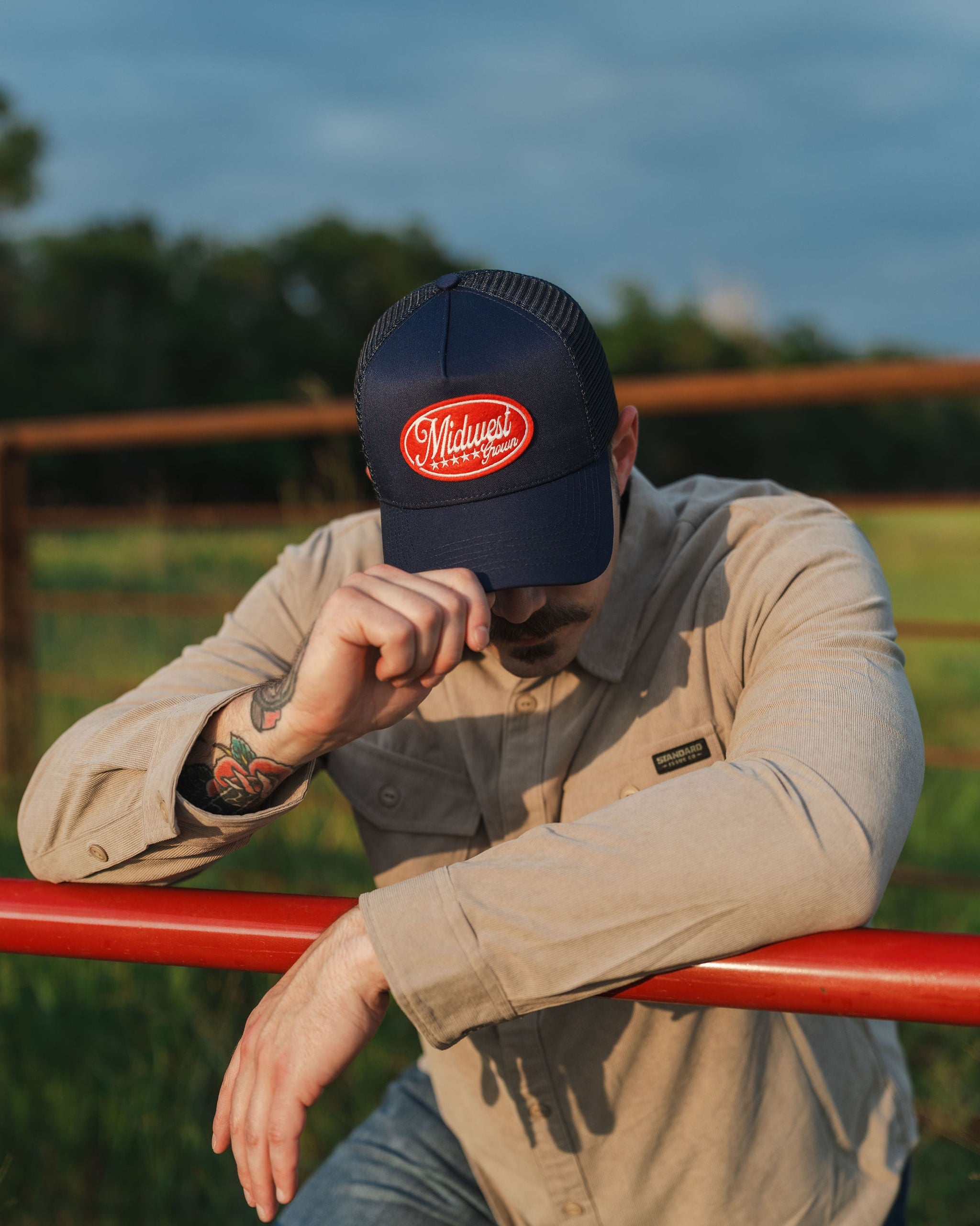 Man wearing a navy cap with a red logo that reads Midwest Grown, sitting on a red fence in an outdoor setting.