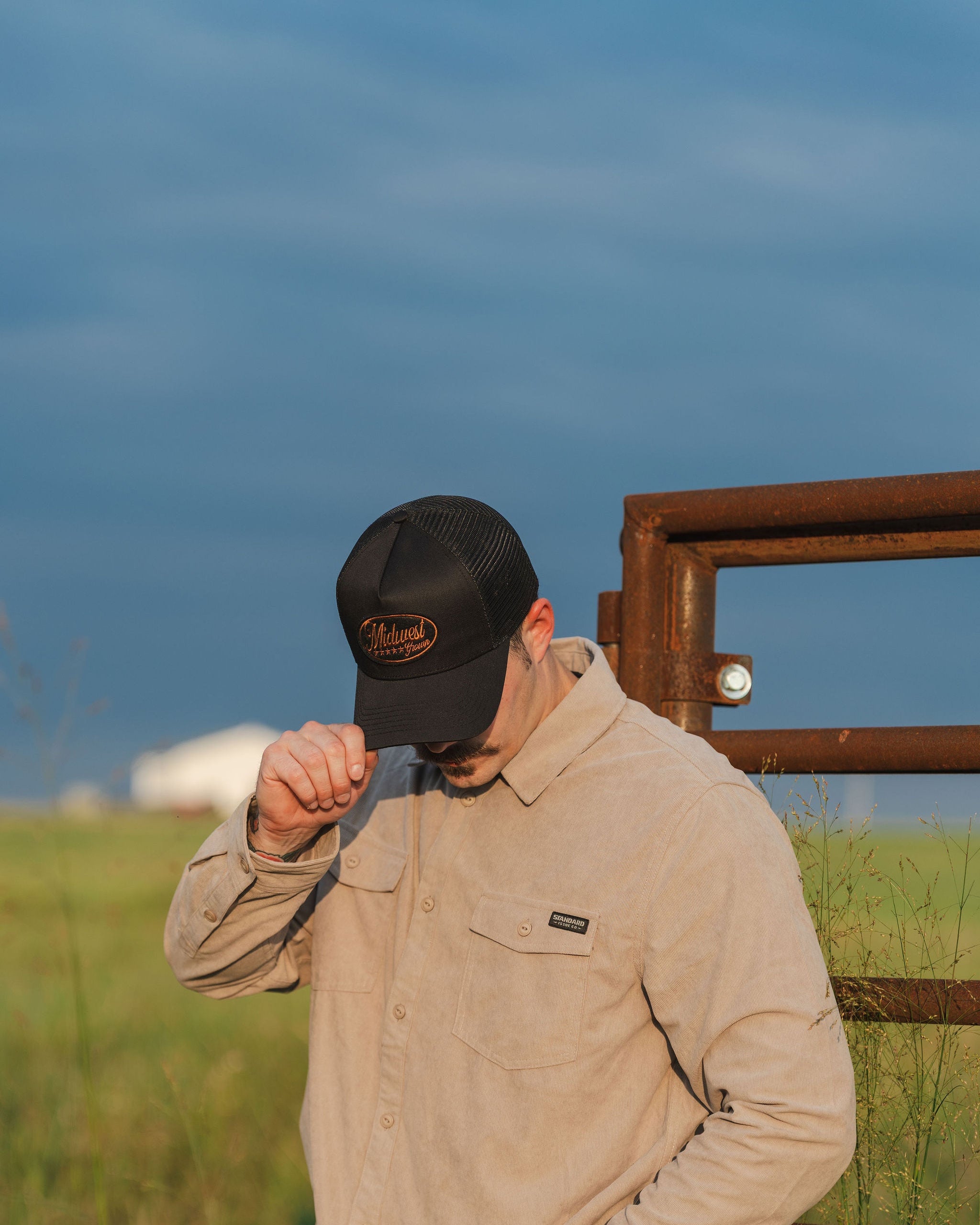 Man wearing a beige jacket and black Midwest Grown cap in a grassy field with a fence and blue sky.