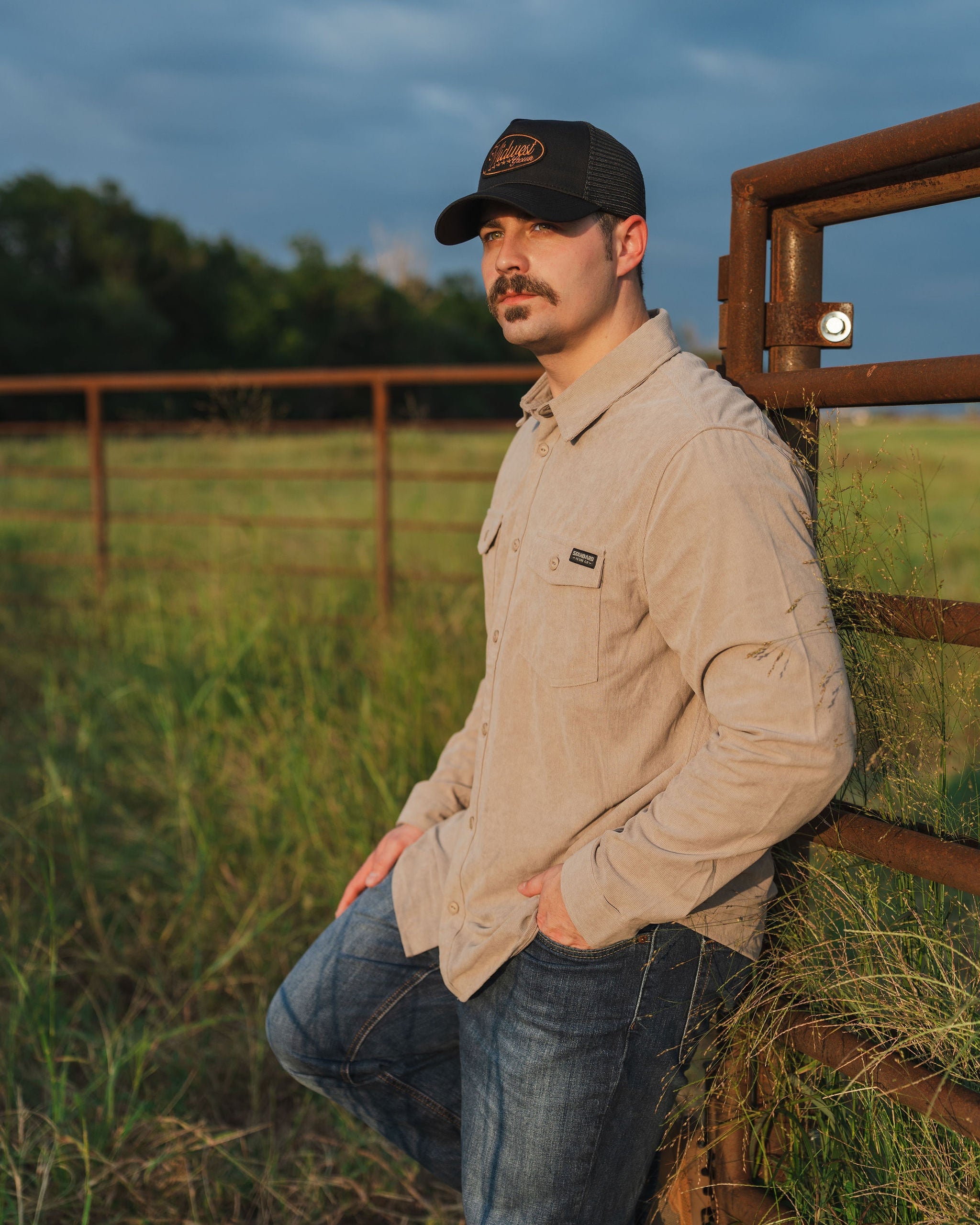 Man leaning against a fence in a field with a dark sky wearing a Midwest Grown hat.