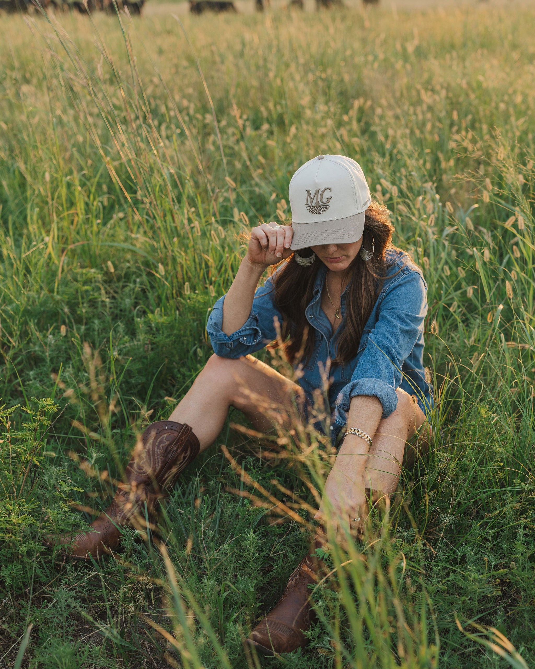 Person sitting in a field wearing a denim jacket and cream Midwest Grown cap.