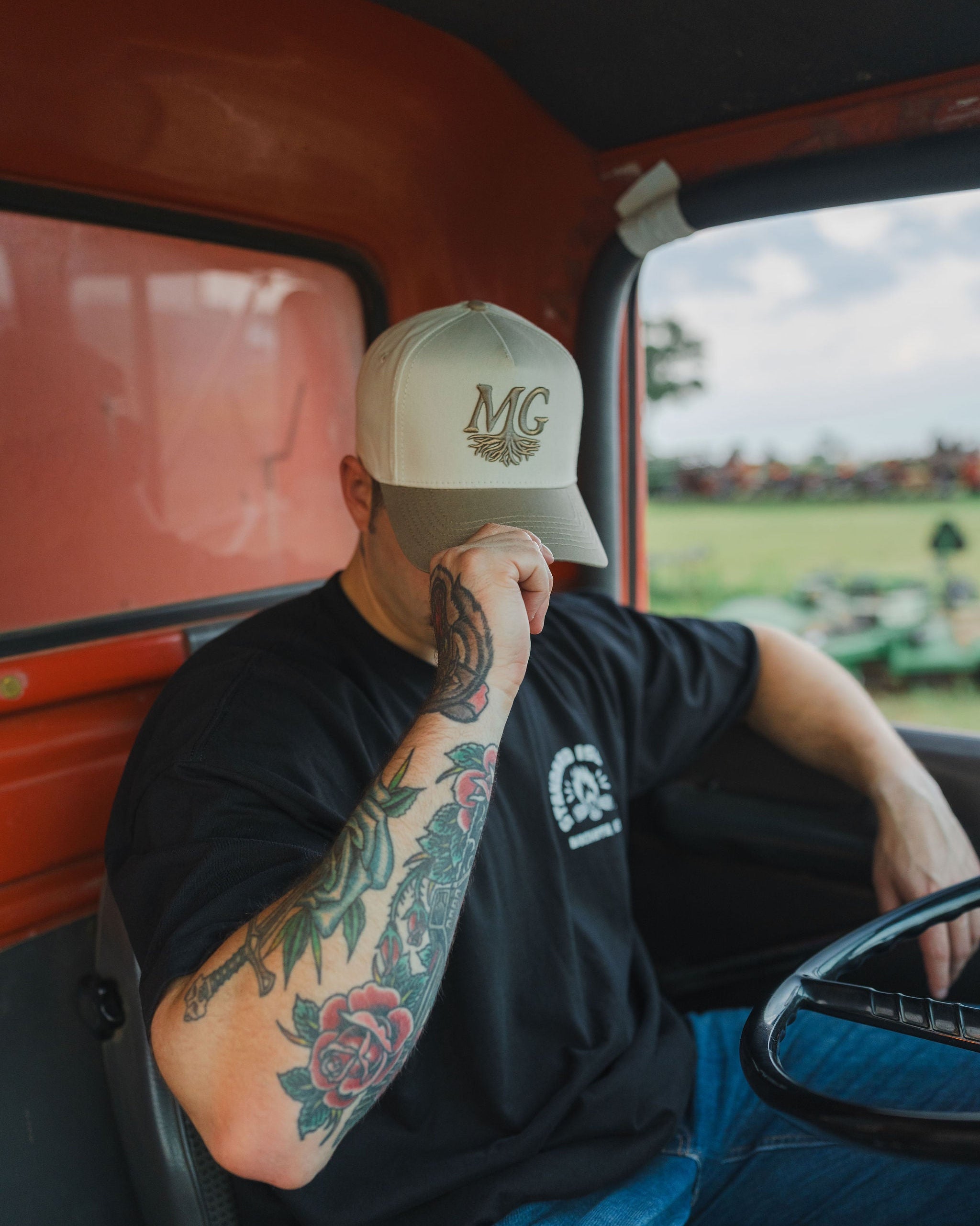 Person sitting inside a vehicle wearing a cap with 'Midwest Grown' logo, tattoos on arm, and a black t-shirt.