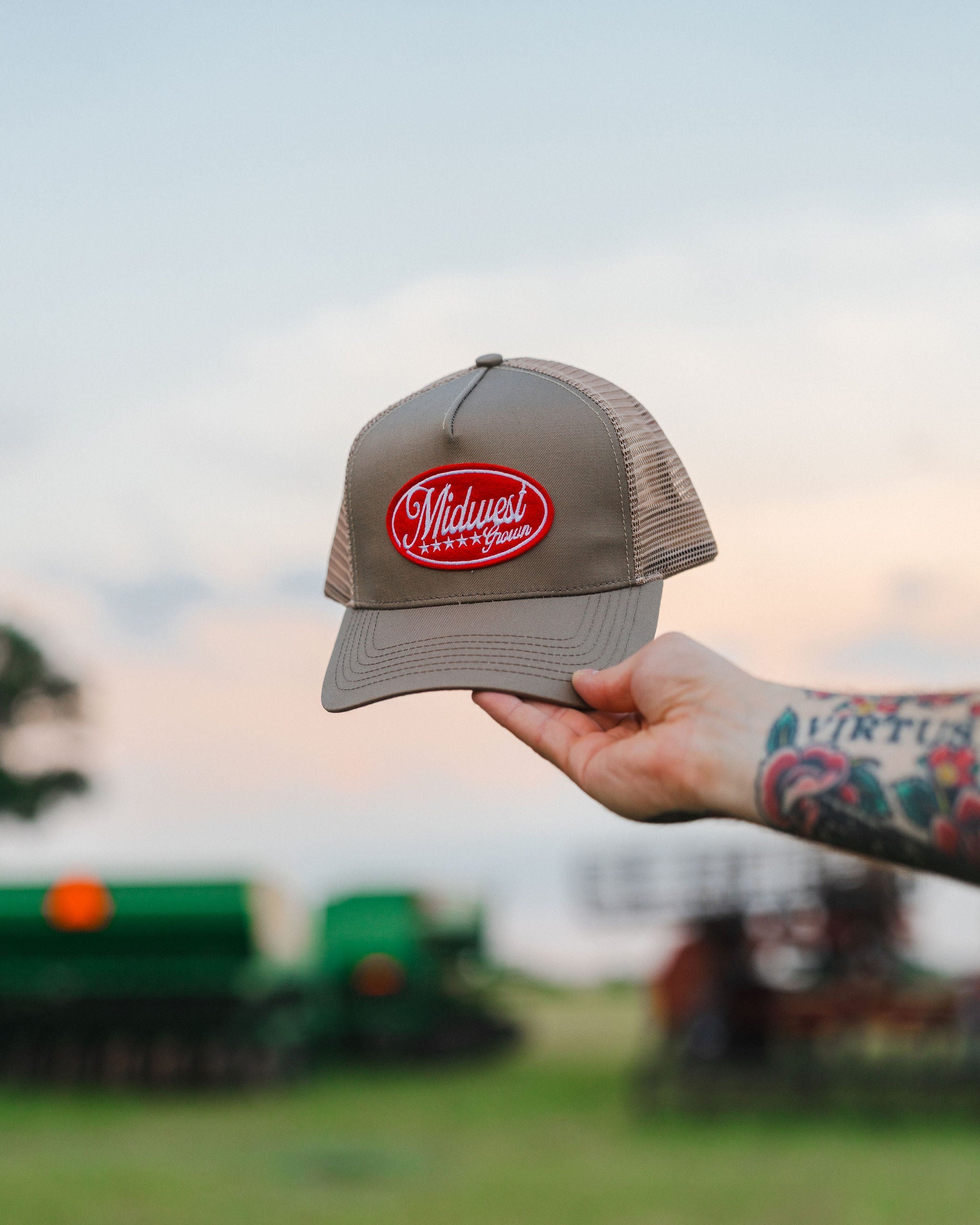 Hand holding a Midwest Grown cap with a logo against a blurred outdoor background