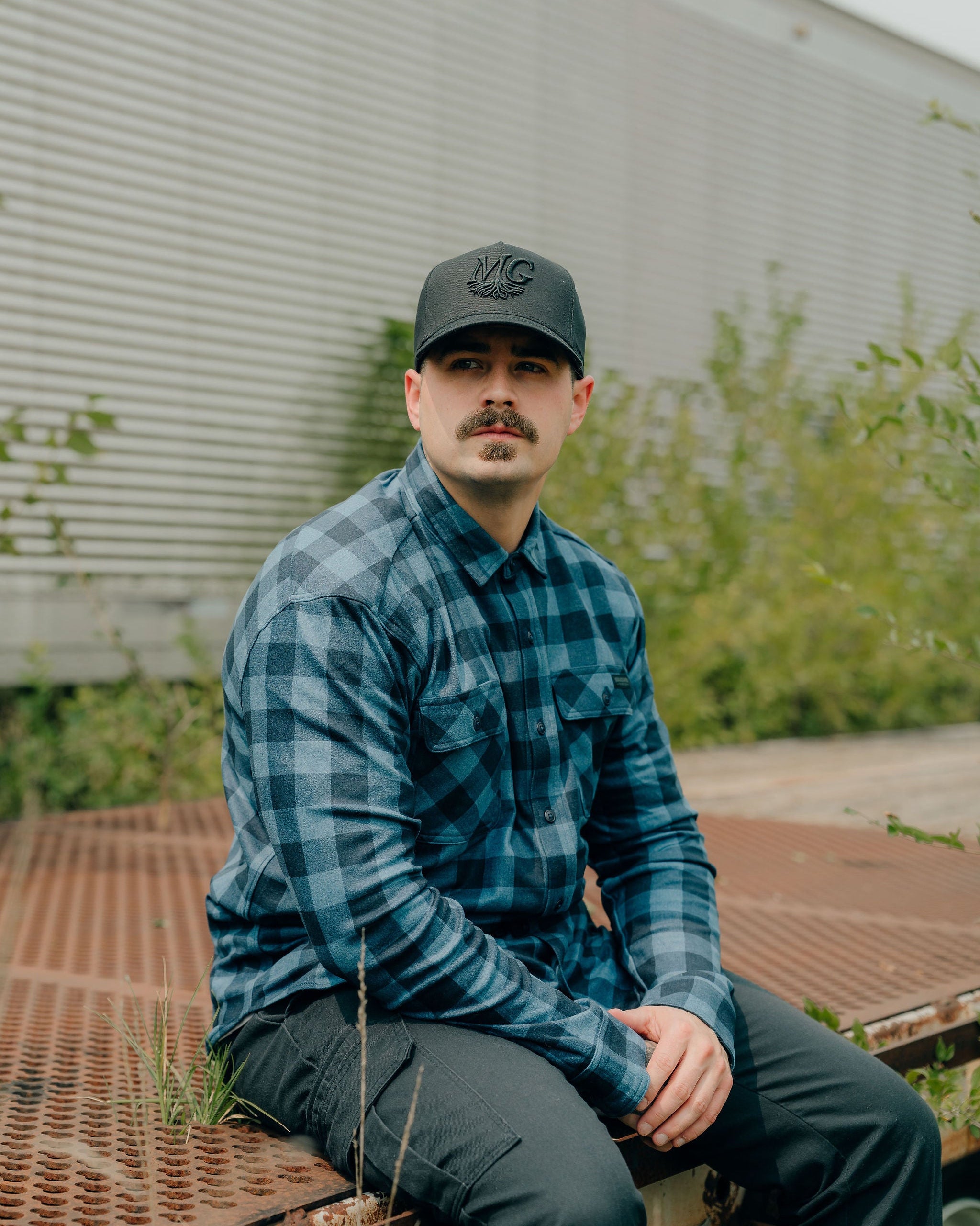Man wearing a blue plaid shirt and a Midwest Grown cap sitting on a metal surface with greenery in the background