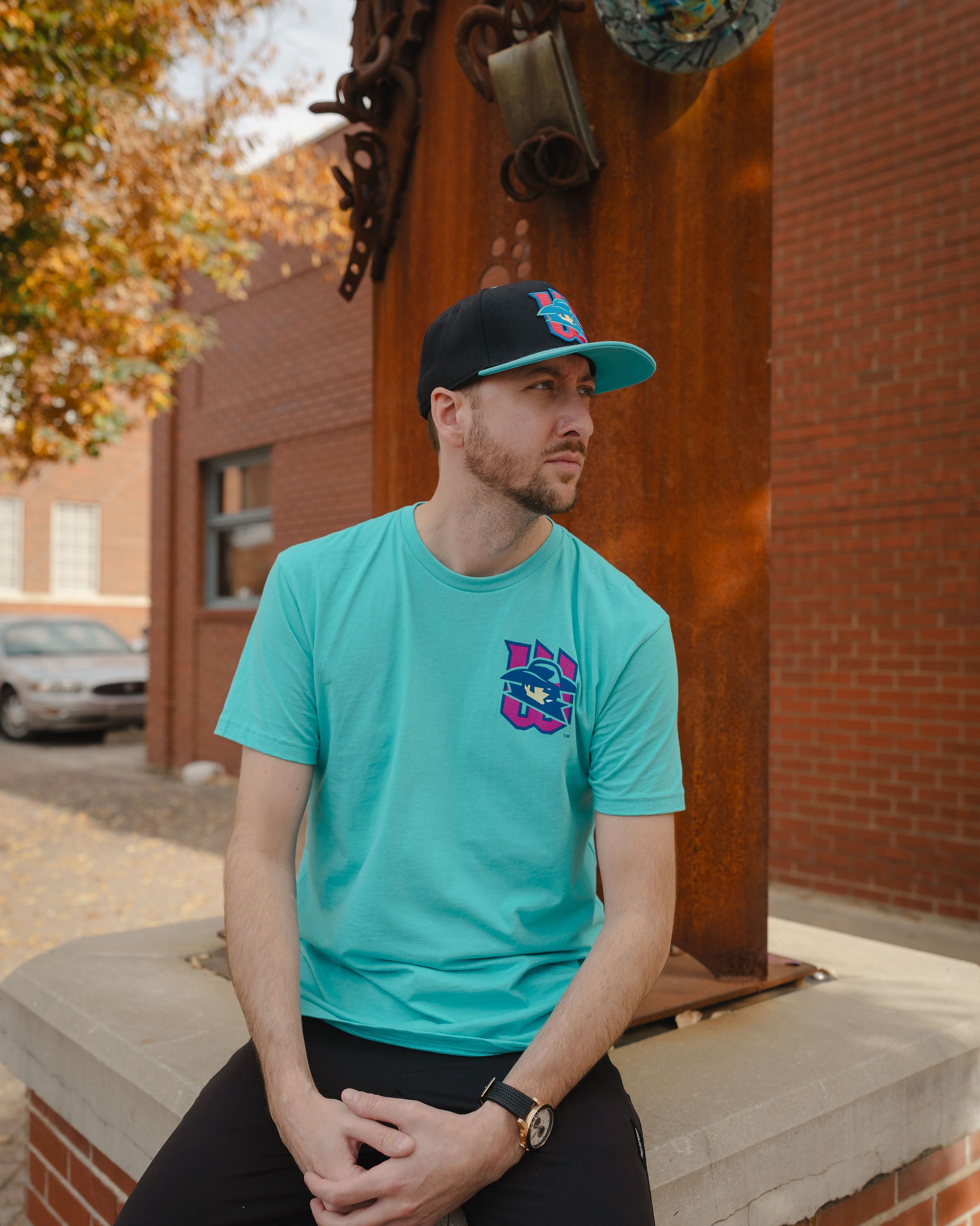Man wearing a teal t-shirt with a logo and a black cap, sitting on a ledge outdoors.