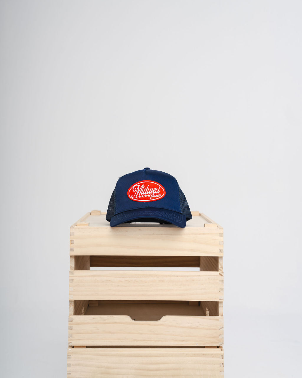Blue Midwest Grown cap with a red logo on a wooden crate against a white background