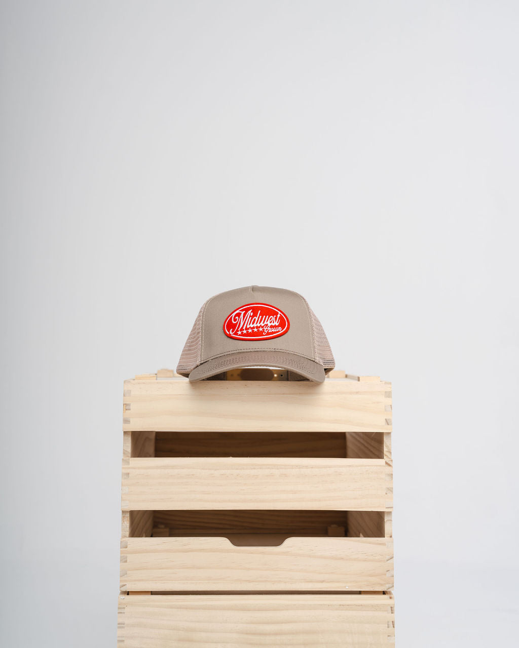 Beige Midwest Grown cap with red logo on a wooden crate against a light gray background