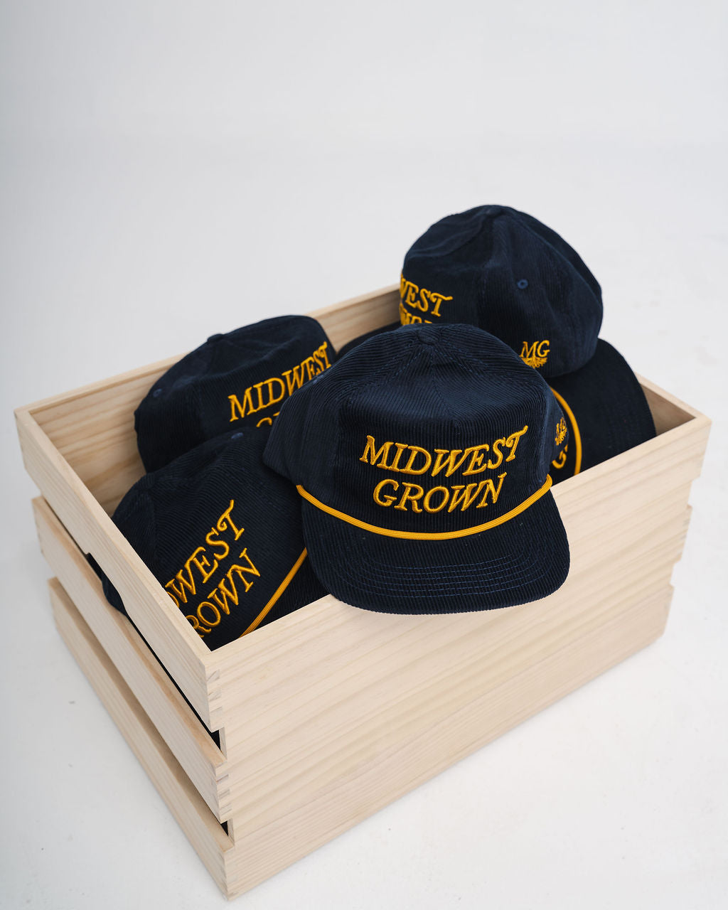 Navy colored hats with 'Midwest Grown' text in yellow on a wooden crate against a white background