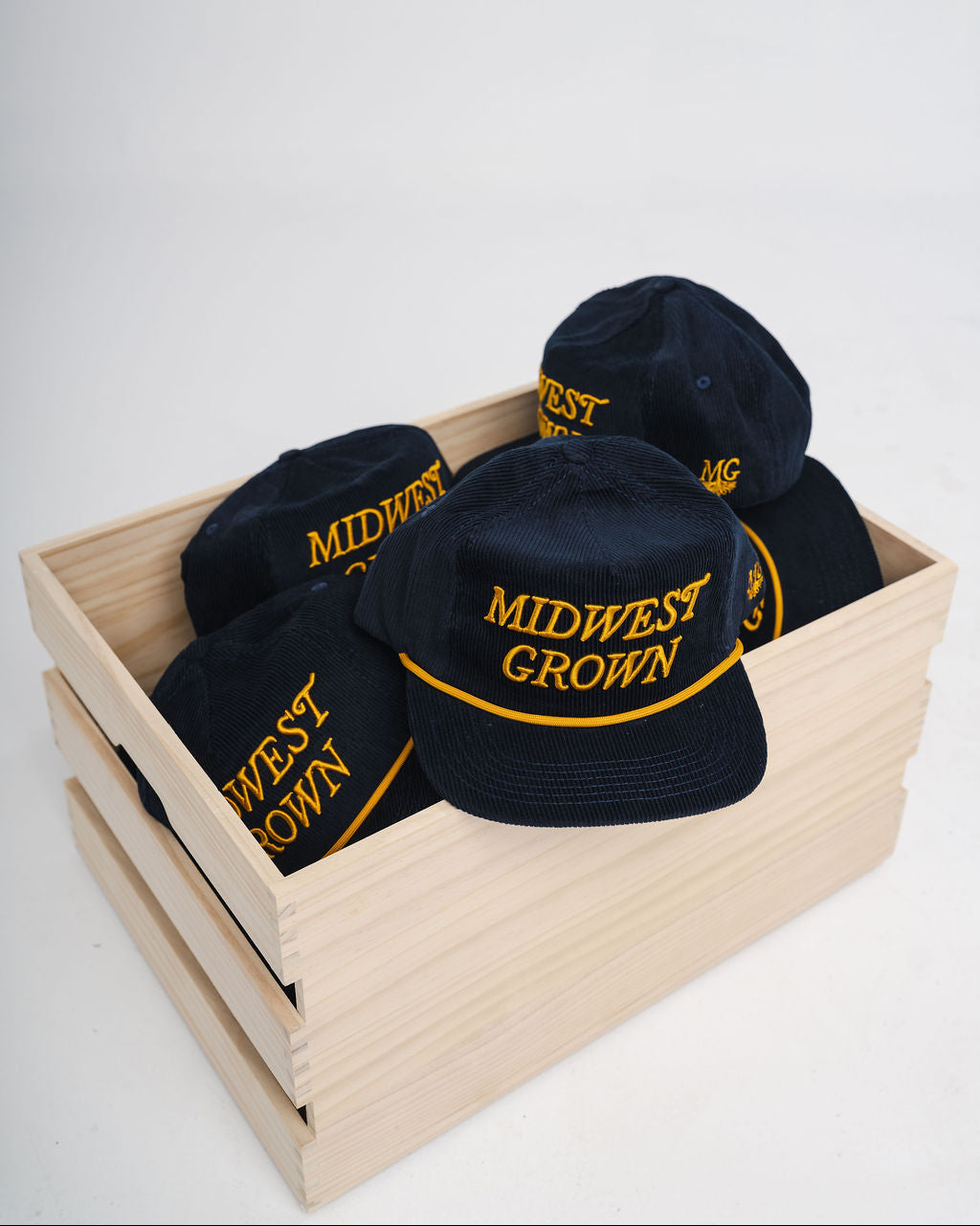 Navy colored hats with 'Midwest Grown' text in yellow on a wooden crate against a white background