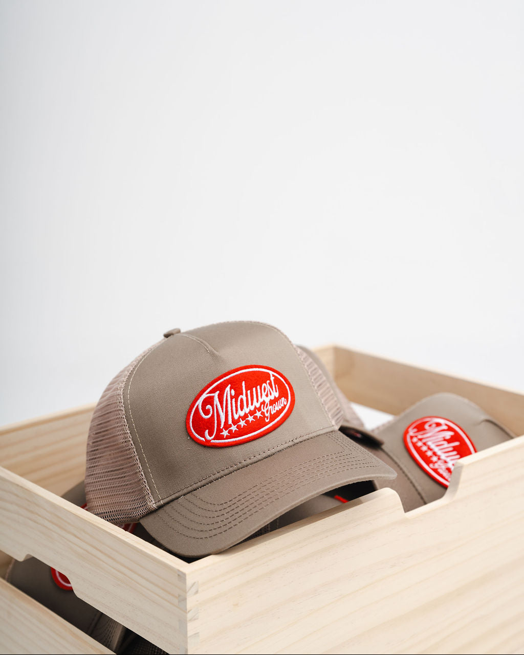 Beige Midwest Grown cap with red logo in a wooden crate on a light gray background