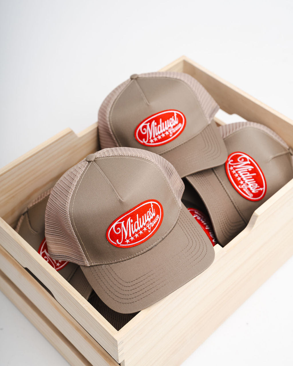 Three beige Midwest Grown caps with red logos in a wooden box on a light gray background