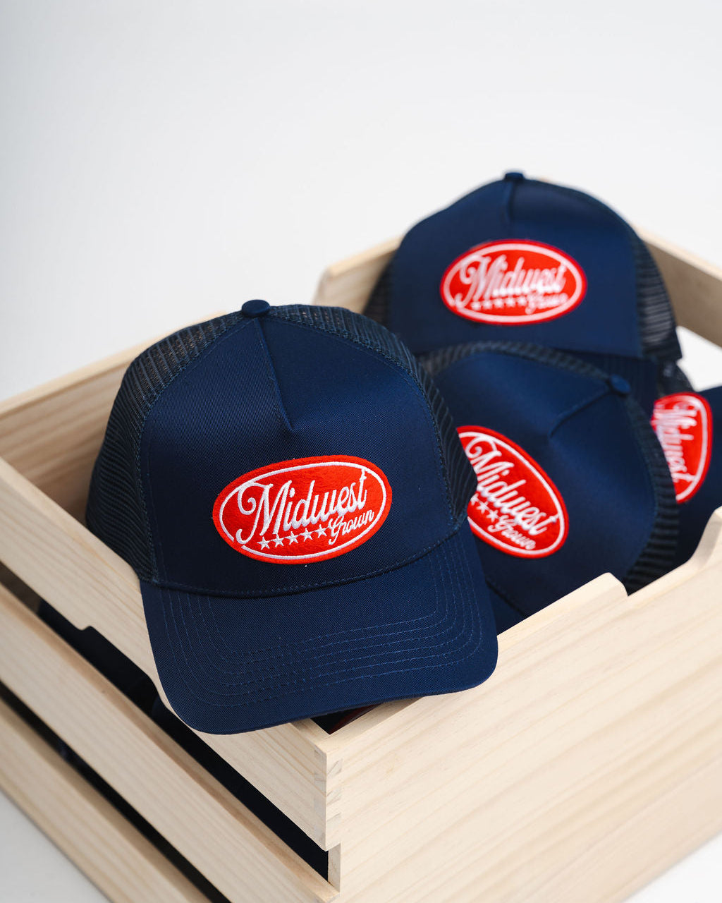 Navy blue caps with red logo reading Midwest Grown in a wooden box on a white background