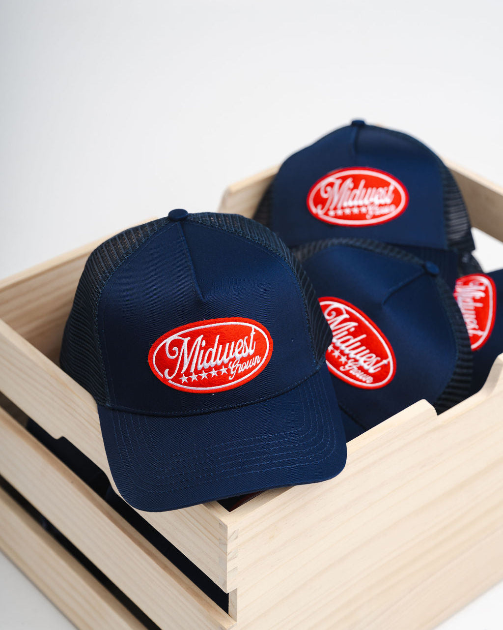 Navy blue caps with red logo reading Midwest Grown in a wooden box on a white background