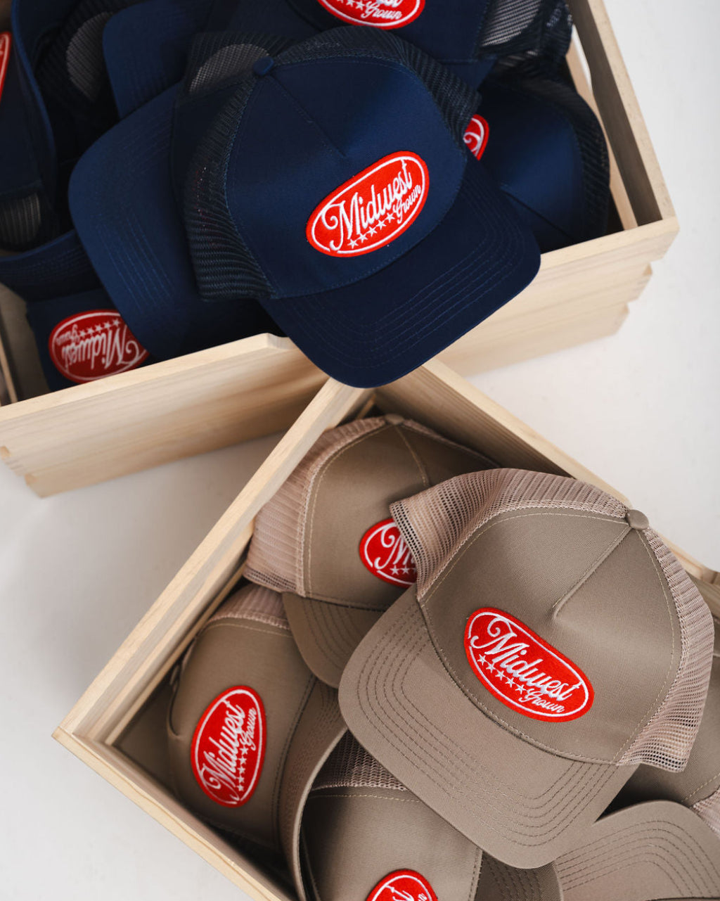 Navy and brown Midwets Grown caps with a red logo in wooden boxes on a white surface