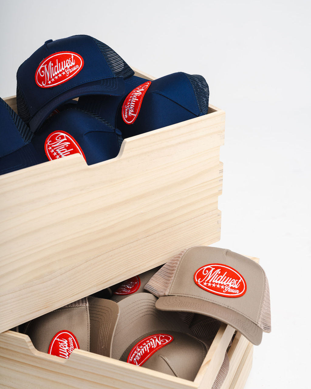 Navy and beige Midwest Grown baseball caps with a red logo in a wooden box on a light gray background