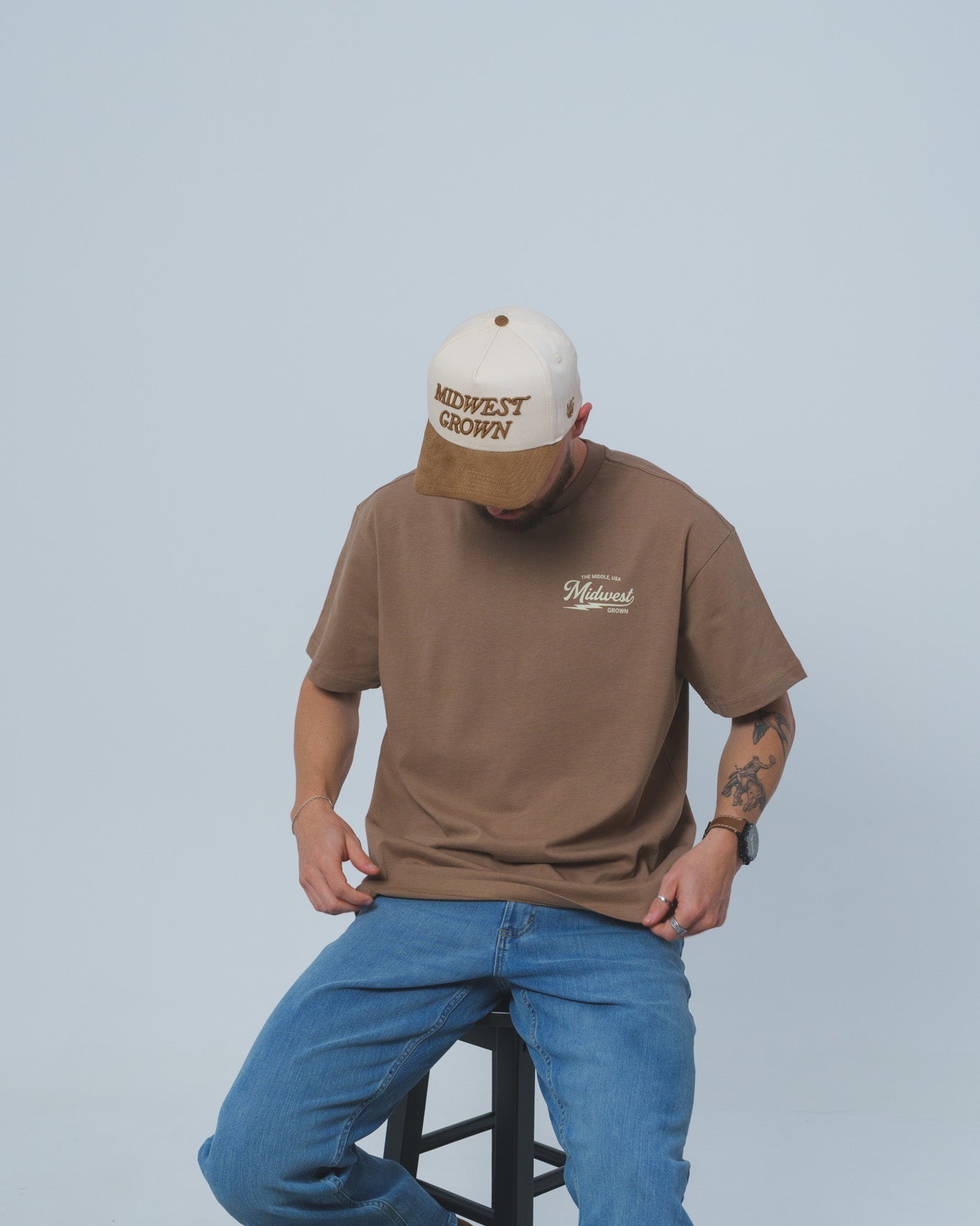 Man wearing a brown Midwest Grown shirt and blue jeans sitting on a stool against a plain background