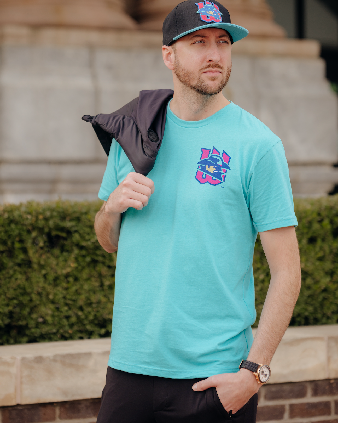Man wearing a teal t-shirt with a colorful logo and a black cap, standing outdoors.