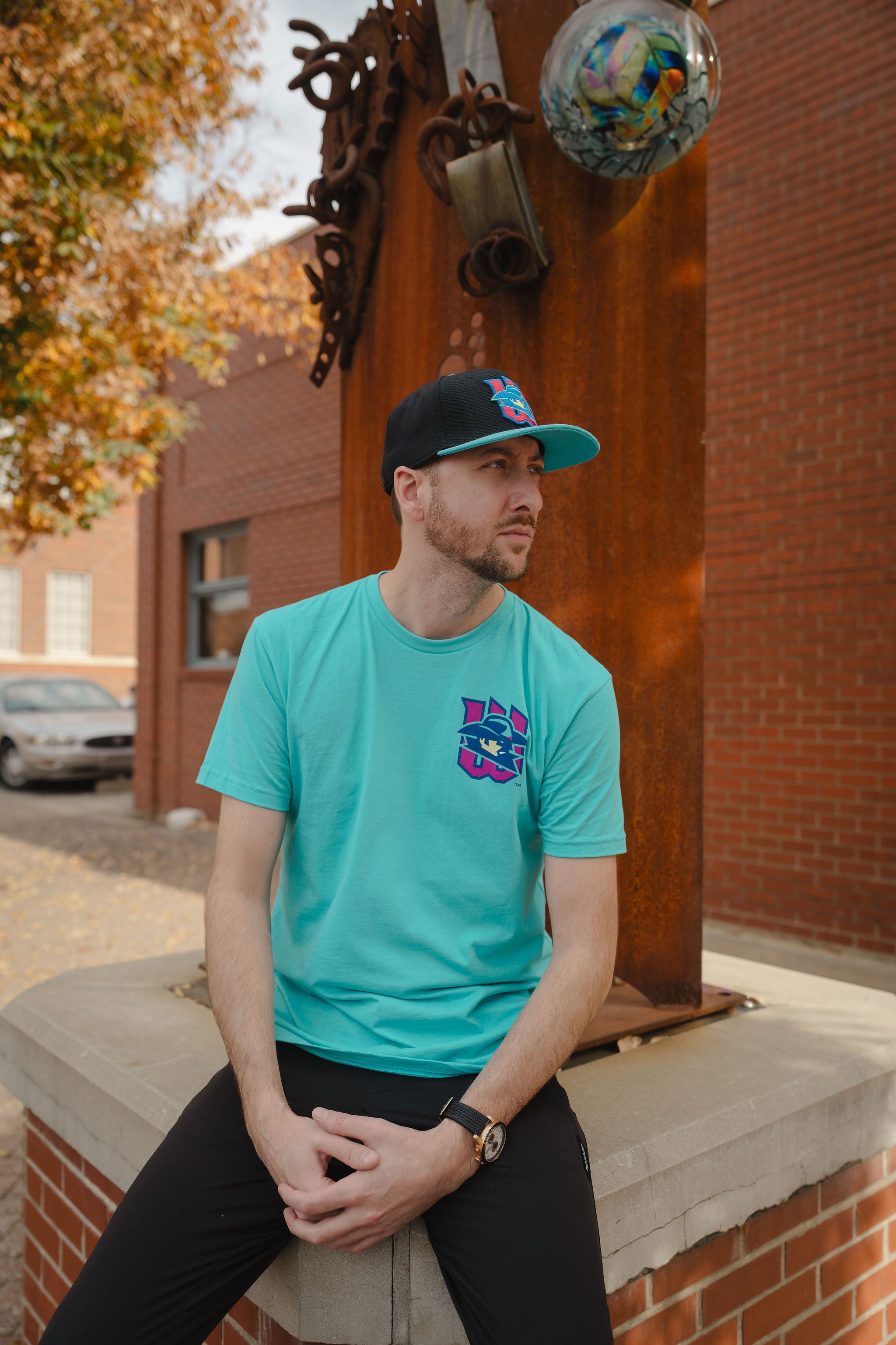 Man wearing a teal t-shirt with a logo and a black cap, sitting on a ledge outdoors.