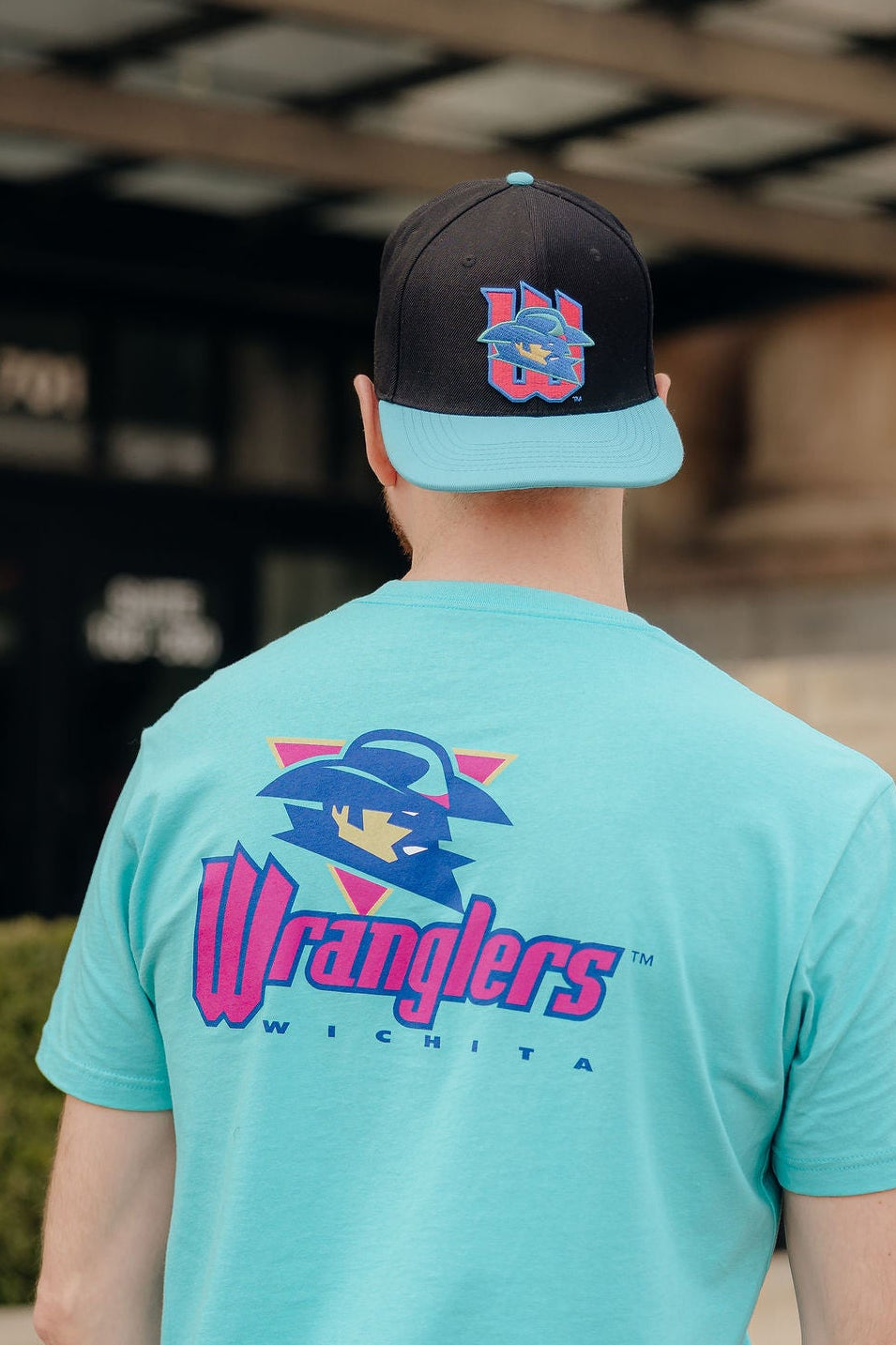 Person wearing a turquoise 'Wranglers' t-shirt and black cap outdoors.