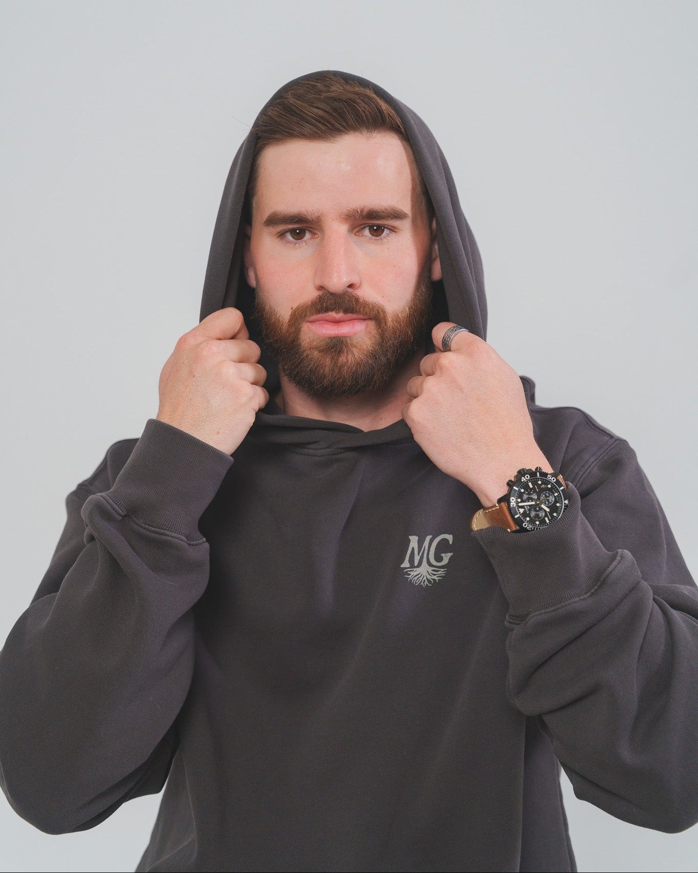 Man wearing a dark hoodie with a Midwest Grown logo, holding the hood with both hands against a light gray background.