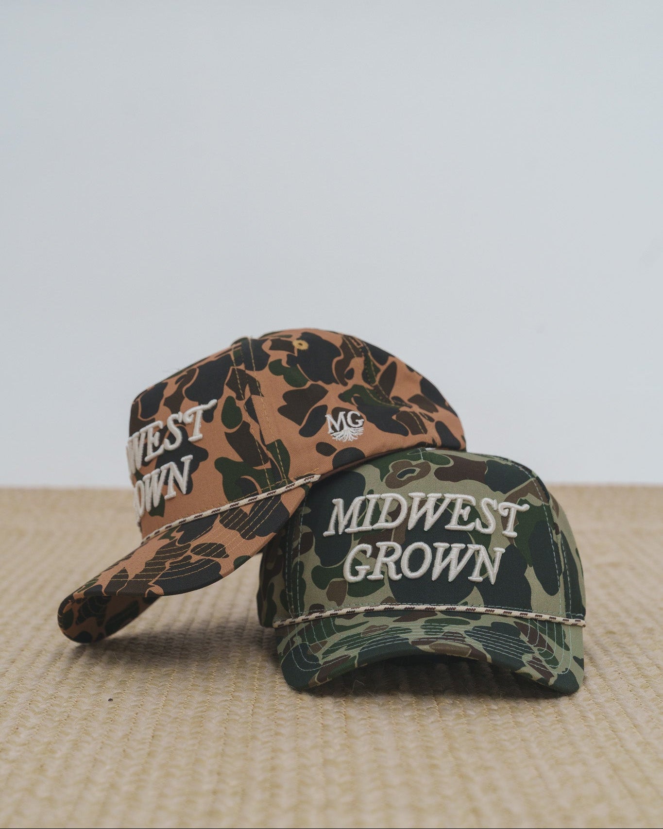Two Duck camouflage baseball caps with 'Midwest Grown' text on a textured surface.