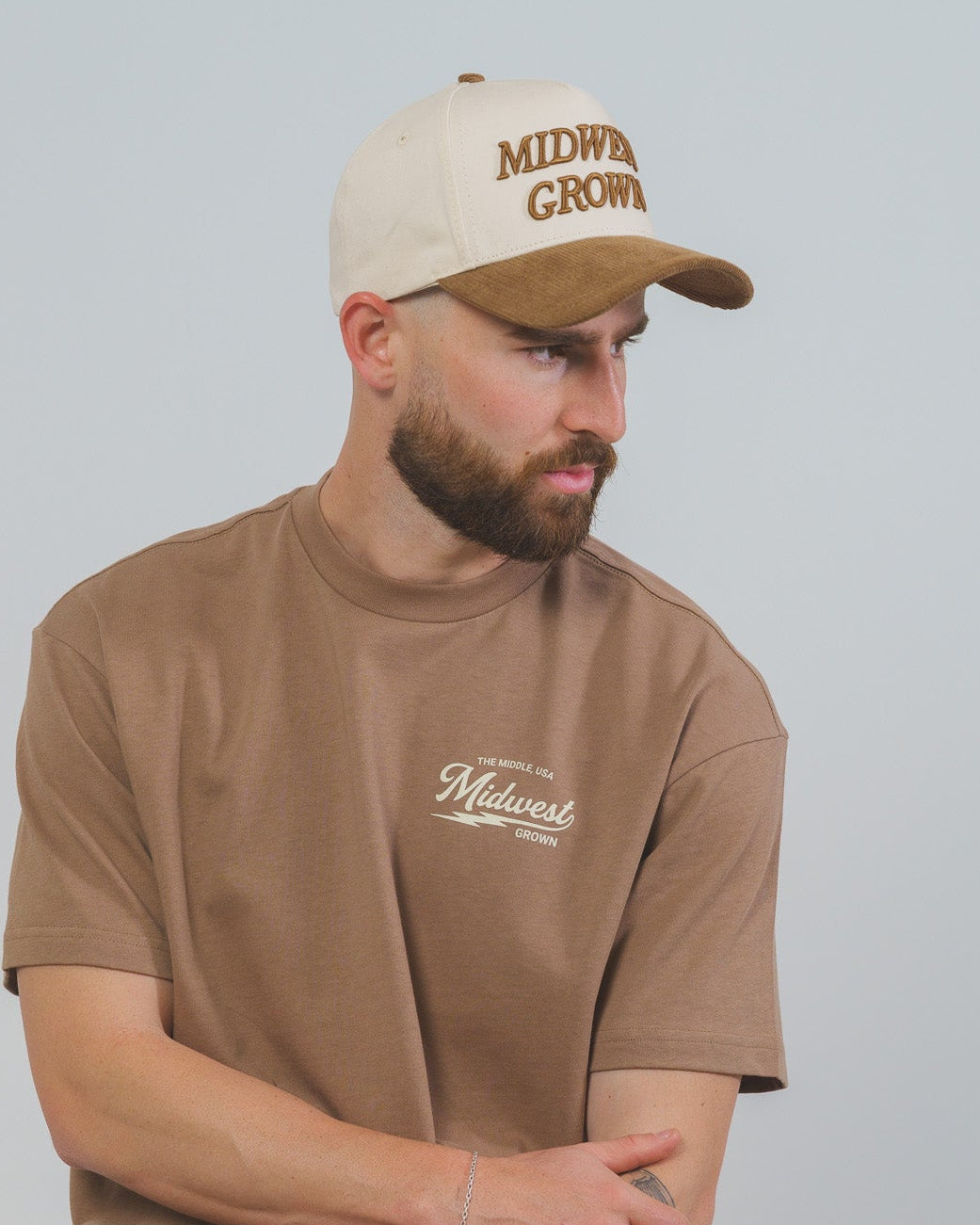 Man wearing a brown t-shirt and cap with 'Midwest Grown' text, sitting on a stool against a plain background.