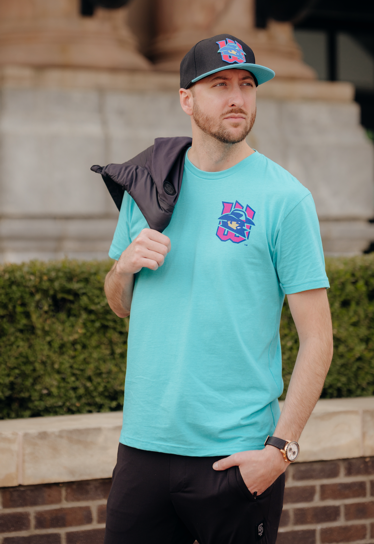Man wearing a teal t-shirt with a colorful logo and a black cap, standing outdoors.